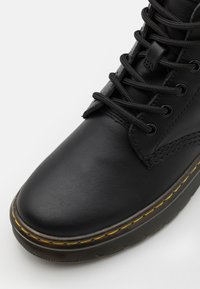 Black leather ankle boot with round toe, thick black laces, and a rugged sole featuring a yellow stitch accent along the edge.