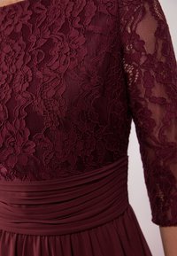 Robe bordeaux avec un corsage en dentelle à motifs floraux, des manches transparentes trois-quarts et une taille froncée pour une apparence texturée.