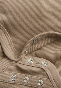 Ribbet beige stof med metalliske tryklåse langs kanten. Blød tekstur, buet halsudskæring og detaljeret syning synlig.