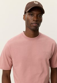 Ροζ βαμβακερό μπλουζάκι με στρογγυλή λαιμόκοψη και κοντά μανίκια, με ανάγλυφη λεπτομέρεια λογότυπου στο αριστερό στήθος. Καφέ μπέιζμπολ καπέλο ολοκληρώνει την εμφάνιση.