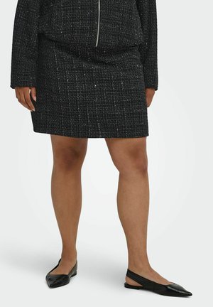 Kvinde iført en sort struktureret nederdel, matchende jakke og sorte spidse slingback-flade sko, stående med benene let adskilt på en hvid baggrund.
