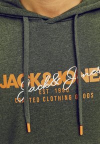 Sudadera de color verde oscuro hecha de tela suave con texto impreso en naranja y blanco. Presenta cordones con puntas naranjas y un diseño clásico de tipo pullover.