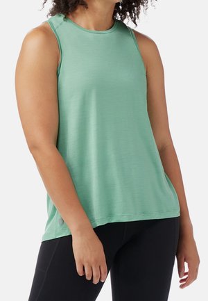 Woman wearing sleeveless light green athletic top and black leggings, standing with arms relaxed at sides against white background.