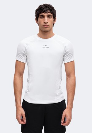 Jeune homme aux cheveux bouclés portant un t-shirt de sport blanc ajusté Nike Pro à manches courtes et un short noir, debout devant un fond uni.
