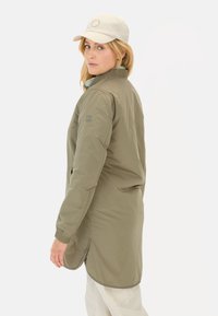 Olijfgroen lange jas met geribbelde kraag, voorzakken en knoopdetail aan de zoom. Draagt een lichte pet en casual broek.