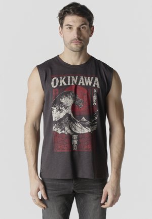 Hombre con cabello corto y oscuro, vistiendo una camiseta negra sin mangas con un gráfico de ola de Okinawa y sol rojo, de pie contra un fondo sencillo.