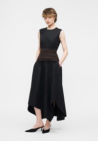 Ženska v ogrnjeni črni obleki brez rokavov z gubanim temno rjavim pasom, asimetričnim robom, rokami v žepih in črnimi špičastimi petami.