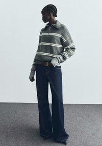 Sivi džemper s bijelim i smeškim prugama, sa zatvaračem na ovratniku i dugim rukavima. Uparen s tamnoplavim širokim farmerkama i smeškim remenom.