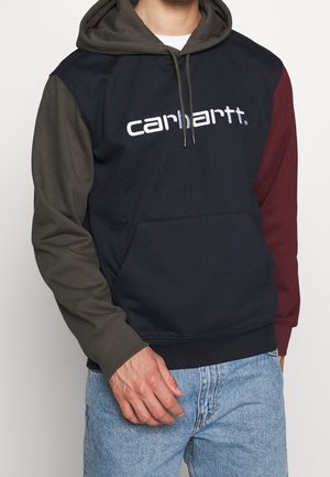 Mand iført en color-block hættetrøje med mørkegrøn ærme og hætte, vinrødt andet ærme, sort krop og hvid "carhartt"-logo på tværs af brystet.