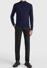 Pull en maille bleu marine à col montant, associé à un pantalon noir ajusté et des chaussures noires, présentant un design simple et une coupe près du corps.