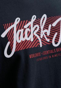 Granatowa bawełniana koszulka z odważnym białym i czerwonym napisem, z graficznymi elementami w paski. Wzór zawiera napis "Jack & Jones" oraz dodatkowy tekst.