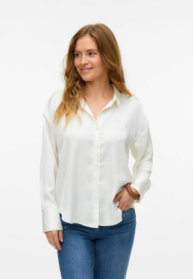 Witte button-up shirt van gladde stof, met een kraag en lange mouwen. Draagt met blauwe denim jeans, relaxed fit, minimale details.