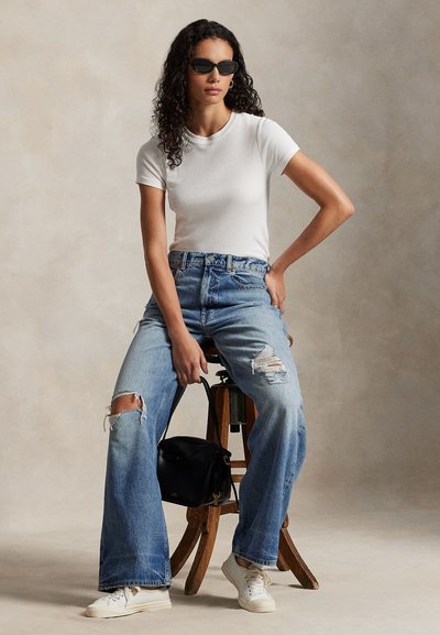 Hvit tettsittende T-skjorte, kombinert med lyseblå, høytlivs, revne jeans. Modellen har på seg svarte solbriller og hvite joggesko, og holder en svart veske.