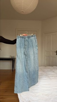 Svetlo modre široke hlače z nežnim leopardjim vzorcem, ki jih na obešalniku drži oseba v minimalistični spalnici z belimi stenami in lesenim podom.