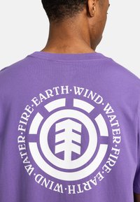 Lilla bomulds t-shirt med et hvidt grafik med en stiliseret træ og cirkulær tekst: "VAND, Ild, JORD, VIND." Bagansigt, crew neck design.
