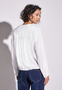 Blusa bianca a maniche lunghe con tessuto texturizzato, vestibilità rilassata e dettaglio arricciato sul retro. Indossata con jeans skinny blu scuro.