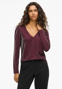 Burgunderrød langermet topp med V-hals, som har en subtil glans og myk tekstur. Ermene har en lett samlet detalj ved skuldrene.
