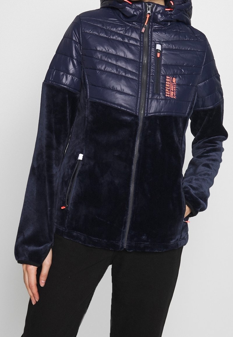 Henkilö, joka on pukeutunut tummansiniseen fleecetakkiin, jossa on tikatut olkapäät, etuvetoketju, vetoketjulliset taskut ja "Superdry"-logo rinnassa.