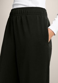 Čierne jogger nohavice s elastickým pásom, hladkou textúrou, bočnými vreckami a voľným strihom, zobrazené na neutrálnom pozadí.