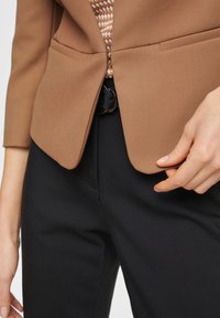 Gros plan sur une personne portant un blazer marron clair, un haut à motifs, un pantalon noir avec une ceinture noire à anneau, ajustant le blazer d'une main.
