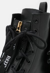 Bottine en cuir noir avec une fermeture éclair dorée, des lacets ronds et des œillets métalliques ; elle présente une texture lisse et une étiquette de marque blanche.