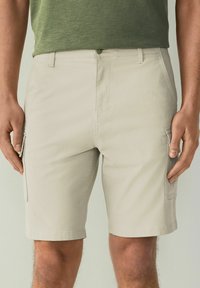Shorts cargo beiges avec poches latérales, fabriqués en tissu léger. Présentent un devant plat, des passants de ceinture et un ourlet droit.