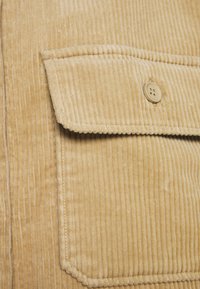 Tejido de pana color marrón con rayas verticales, que presenta un bolsillo con solapa y un detalle de botón. Textura suave y ribeteado prominente en toda su extensión.