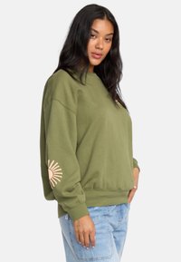Felpa verde con scollo rotondo e maniche lunghe. Caratterizzata da una grafica di sole pesca sulla manica sinistra. Abbinata a jeans blu chiaro.