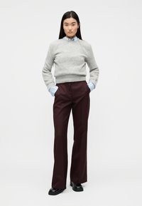 Γυναίκα με γκρι πουλόβερ πάνω από μπλε πουκάμισο, μπορντό παντελόνα με φαρδιά μπατζάκια και μαύρα παπούτσια, όρθια με τα χέρια στις τσέπες σε λευκό φόντο.