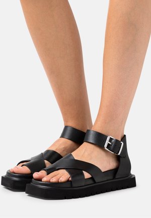 Sandales en cuir noir avec des straps croisées et une boucle à la cheville. Semelle épaisse avec une surface texturée qui ajoute de la stabilité.