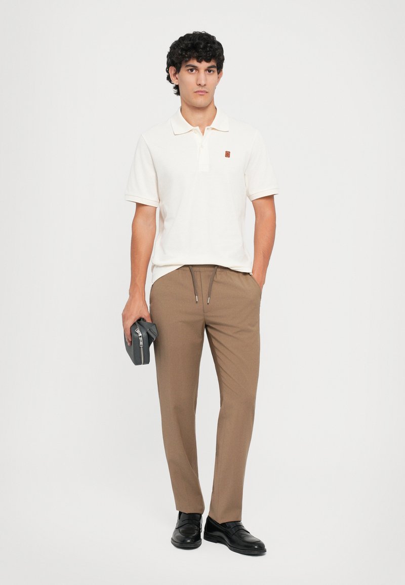 Herren Polo-Shirt in hellbeige mit kurzen Ärmeln und gesticktem Logo, kombiniert mit braunen Hosen und schwarzen Dressschuhen. Hält ein graues Accessoire.