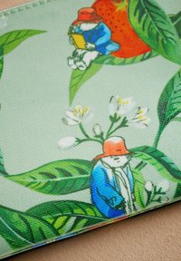 Mønstrede stof har grønne blade, en kanin i en blå frakke med en rød hat, og blomster, alt sammen sat mod en lys grøn baggrund.