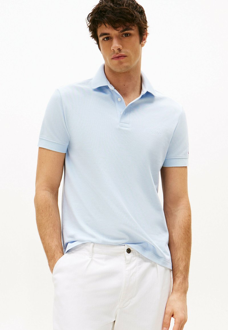 Polo shirt azzurro chiaro con maniche corte, colletto e un logo sottile. Indossato con pantaloni bianchi, che mostrano un design casual e aderente.