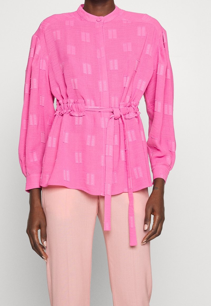 Blusa rosa con un motivo testurizzato, stretta in vita da una cintura. Le maniche sono a palloncino e presentano un colletto alto.