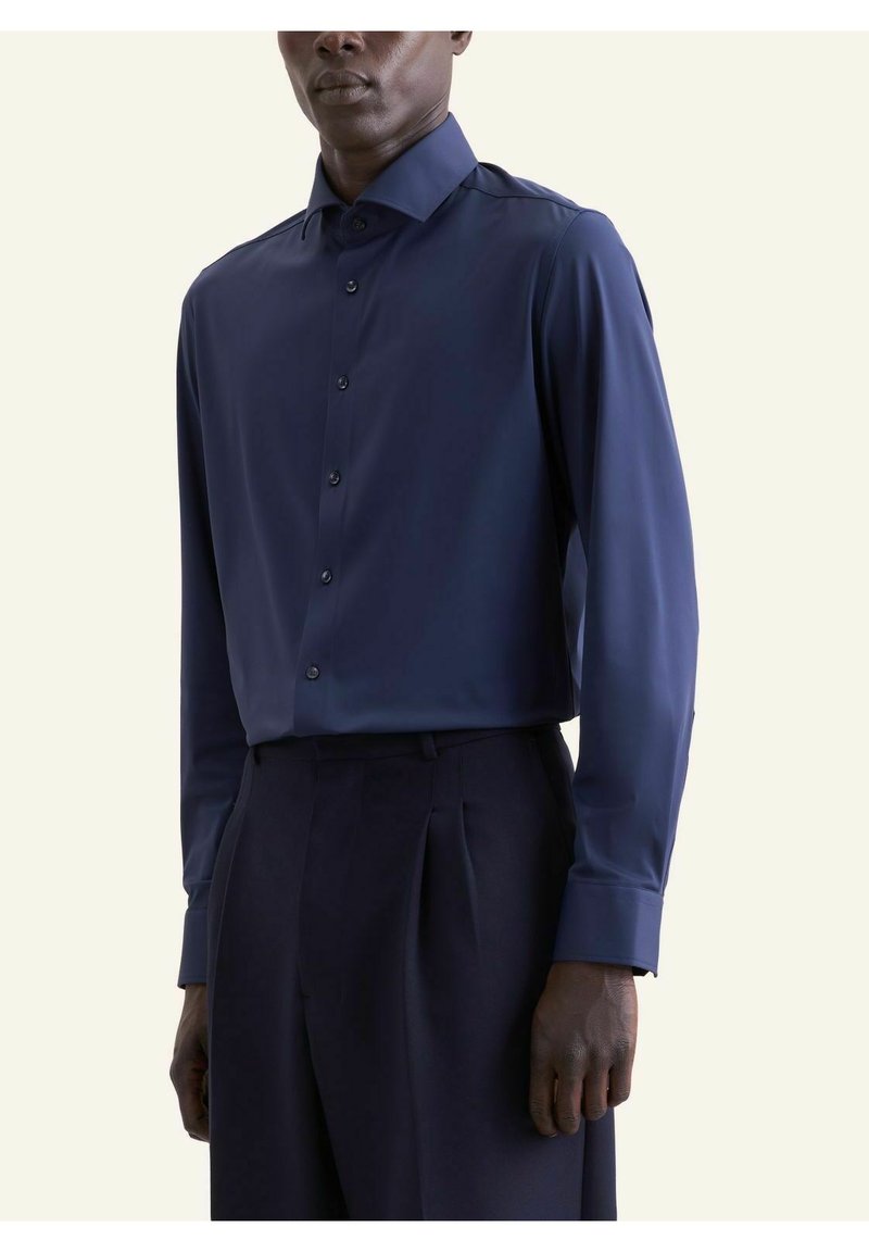 Homme portant une chemise à manches longues bleu marine boutonnée, rentrée dans un pantalon plissé bleu marine taille haute, sur un fond uni.