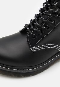 Bottine en cuir noir avec des coutures blanches, six œillets, lacets noirs avec des accents blancs, bout arrondi et design de semelle robuste.