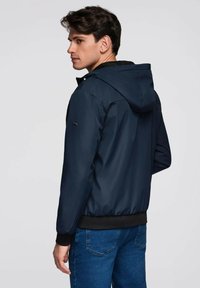 Mladý muž v námořnické kapucové bundě a modrých džínách, stojící nalevo s neutrálním výrazem na pozadí v světlé šedé barvě.