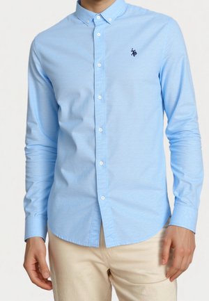 Mann trägt ein hellblaues, langärmliges Hemd mit Knöpfen und beige Hosen, steht vor einem schlichten Hintergrund.