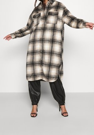Woman wearing oversized beige and black plaid button-up shirt dress, black gathered faux leather pants, and black strappy heels.