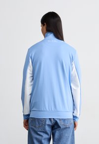 Light blue full-zip jacket with high collar and white long sleeves. Made from smooth, stretchy fabric with minimal detailing on the back.