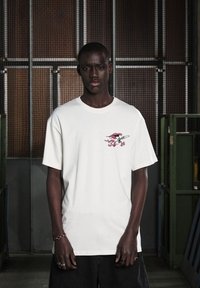 Camiseta de algodón blanca con un gráfico colorido en el pecho izquierdo. Presenta un corte relajado con mangas cortas y un cuello redondo.