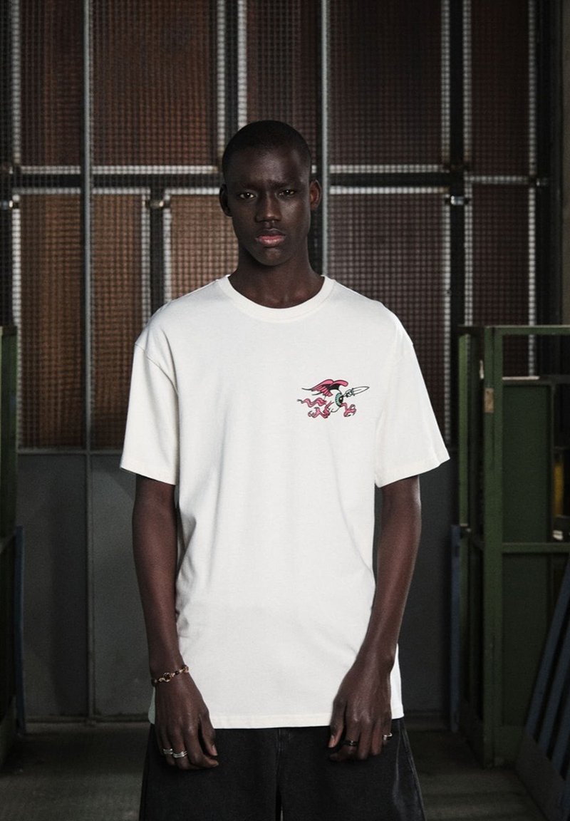 Camiseta de algodón blanca con un gráfico colorido en el pecho izquierdo. Presenta un corte relajado con mangas cortas y un cuello redondo.