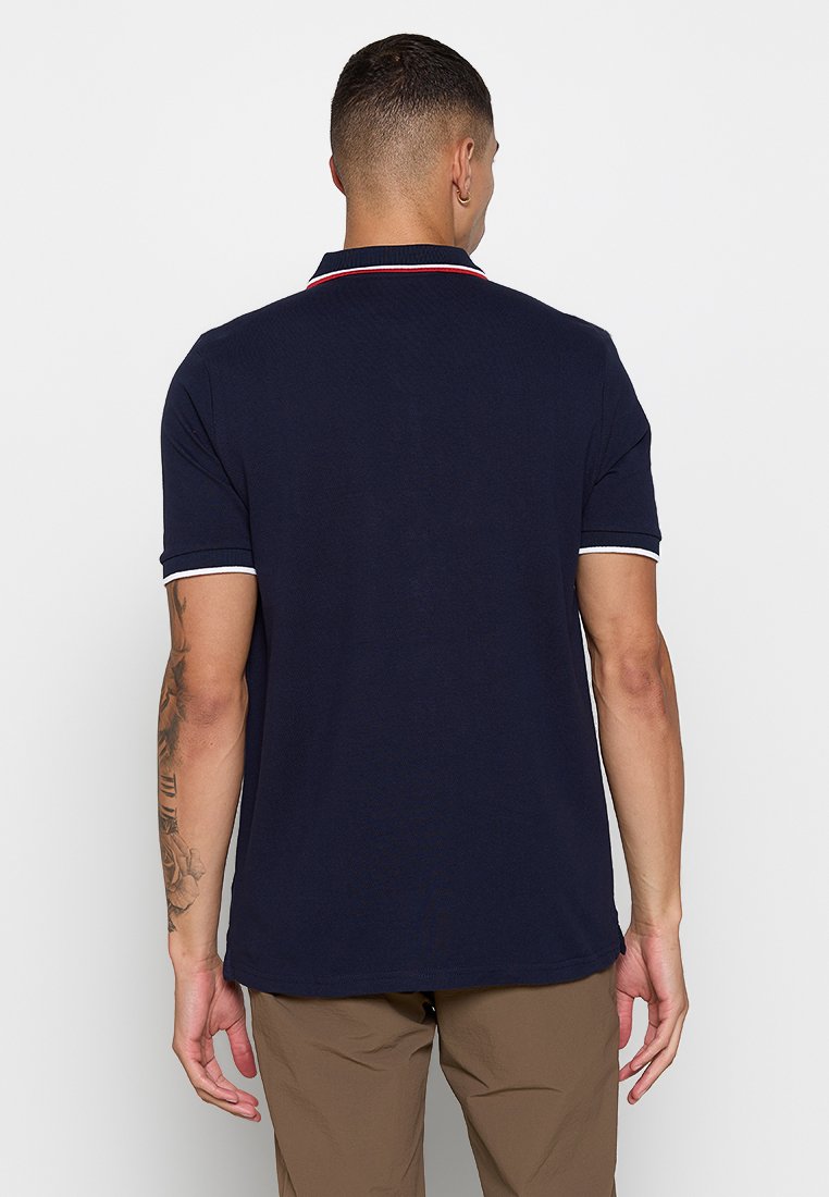 Homme aux cheveux courts portant un polo bleu marine avec des bordures blanches et rouges, un pantalon marron, et montrant un tatouage sur son bras gauche, vu de dos.