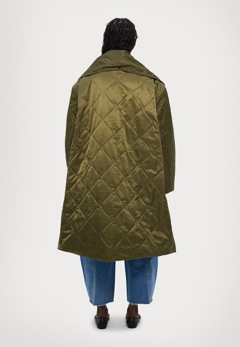 Olivgrüner, gesteppten langer Mantel mit breitem Kragen und Rückenbelüftung, kombiniert mit blauen weit geschnittenen Jeans und schwarzen Mules. Glatte, glänzende Textur.