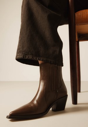 Femeie purtând ghete joase din piele maro cu vârfuri ascuțite și toc bloc, așezată pe un scaun de lemn, cu pantaloni maro care-i acoperă gleznele.