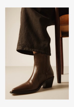 Naine, kellel on jalas pruunid nahast pahkluu saapad teravate varvaste ja plokktaldadega, istub puidust toolil, pruunide pükstega, mis katavad pahkluud.