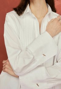 Chemise blanche boutonnée avec devant plissé, manches longues et boutons de manchette dorés ornés d'un motif décoratif d'insecte sur chaque poignet.