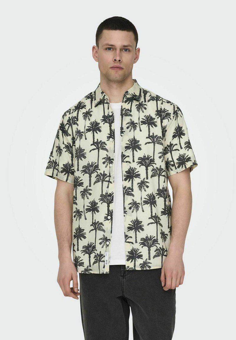 Chemise à manches courtes de couleur jaune clair avec un motif de palmier noir. Elle présente un design à boutons et une coupe décontractée. Portée par-dessus un t-shirt blanc.