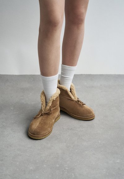 Botas de tornozelo em camurça castanha com forro de pele de ovelha, apresentando um bico arredondado, atacadores e uma sola de borracha, combinadas com meias brancas com nervuras.