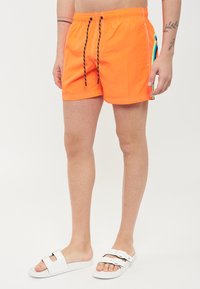 Costumi da bagno arancione brillante realizzati in materiale leggero, con vita regolabile, strisce laterali a contrasto e dettagli di cucitura bianca.